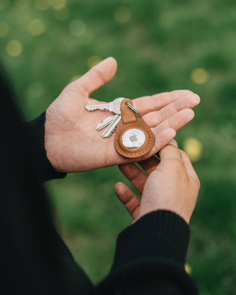 Hand holding a set of keys with an Apple AirTag keychain against a blurred green background